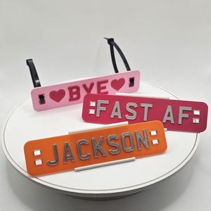 May include: Four rectangular luggage tags in pink, red, and orange. The tags have black straps and feature the phrases "BYE", "FAST AF", and the name "JACKSON" in silver lettering. The tag with "BYE" has a heart symbol.