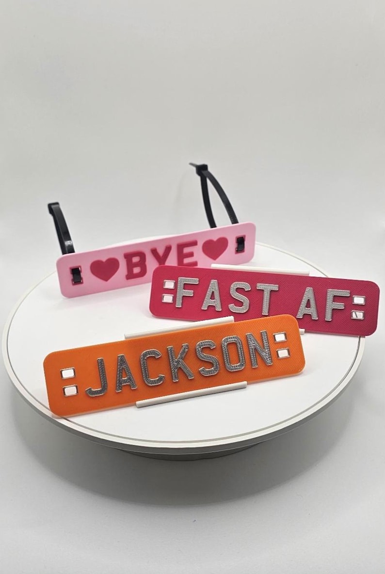 May include: Four rectangular luggage tags in pink, red, and orange. The tags have black straps and feature the phrases "BYE", "FAST AF", and the name "JACKSON" in silver lettering. The tag with "BYE" has a heart symbol.