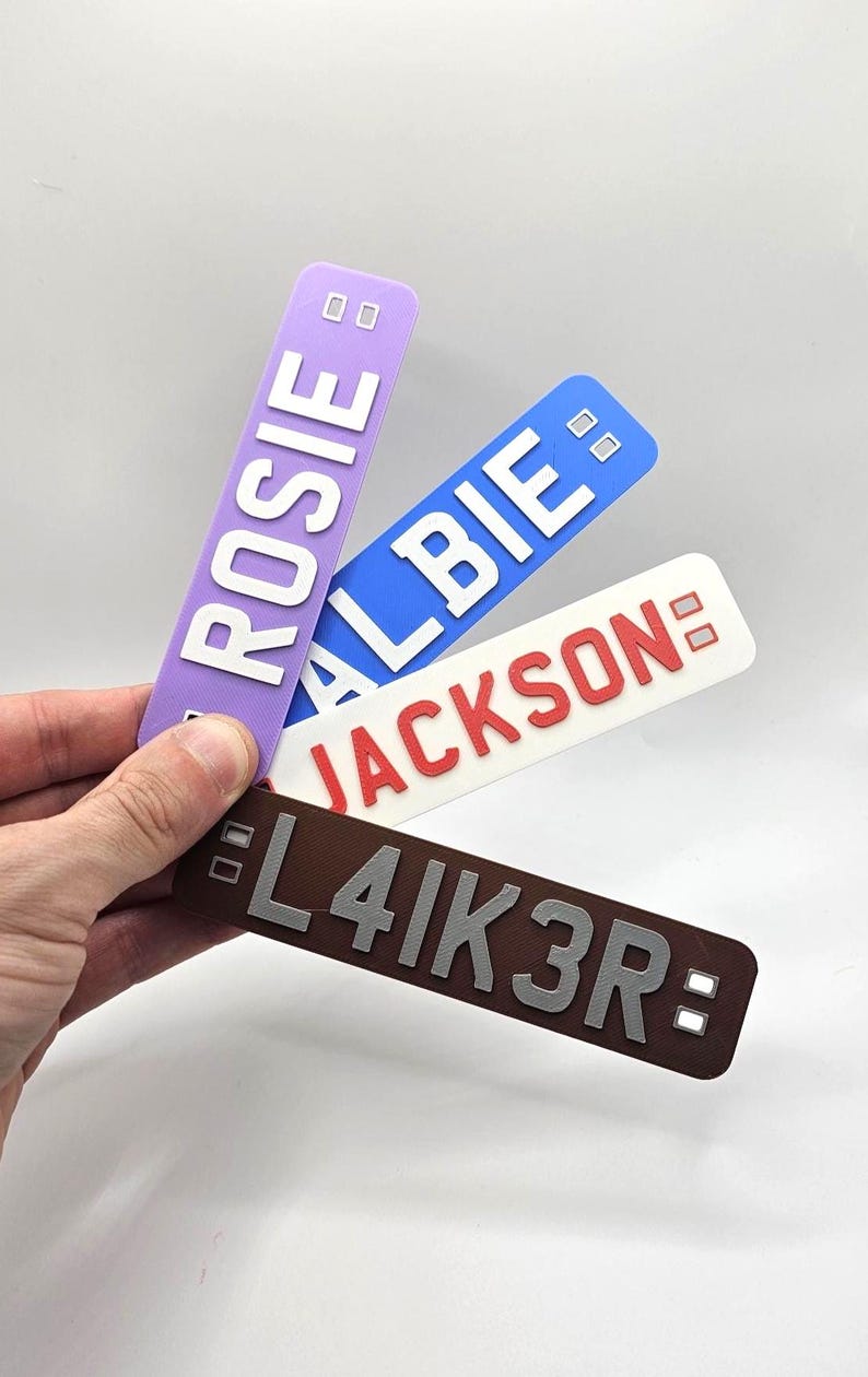 May include: A hand holds four rectangular nameplates in various colors: purple, blue, white, and brown. Each plate features a different name or word in bold, white or silver lettering: "ROSIE", "ALBIE", "JACKSON", and "L4IK3R:".