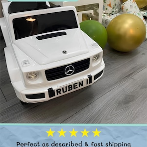 May include: A white toy car with black accents, featuring a front grille with the Mercedes-Benz logo and the name "RUBEN" on the license plate. The text "Small Plate Example" is at the top. A gold and green balloon are in the background.