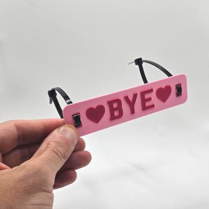 May include: A pink luggage tag with the word "BYE" and heart symbols in dark pink. The tag is held by black zip ties. The background is plain white.