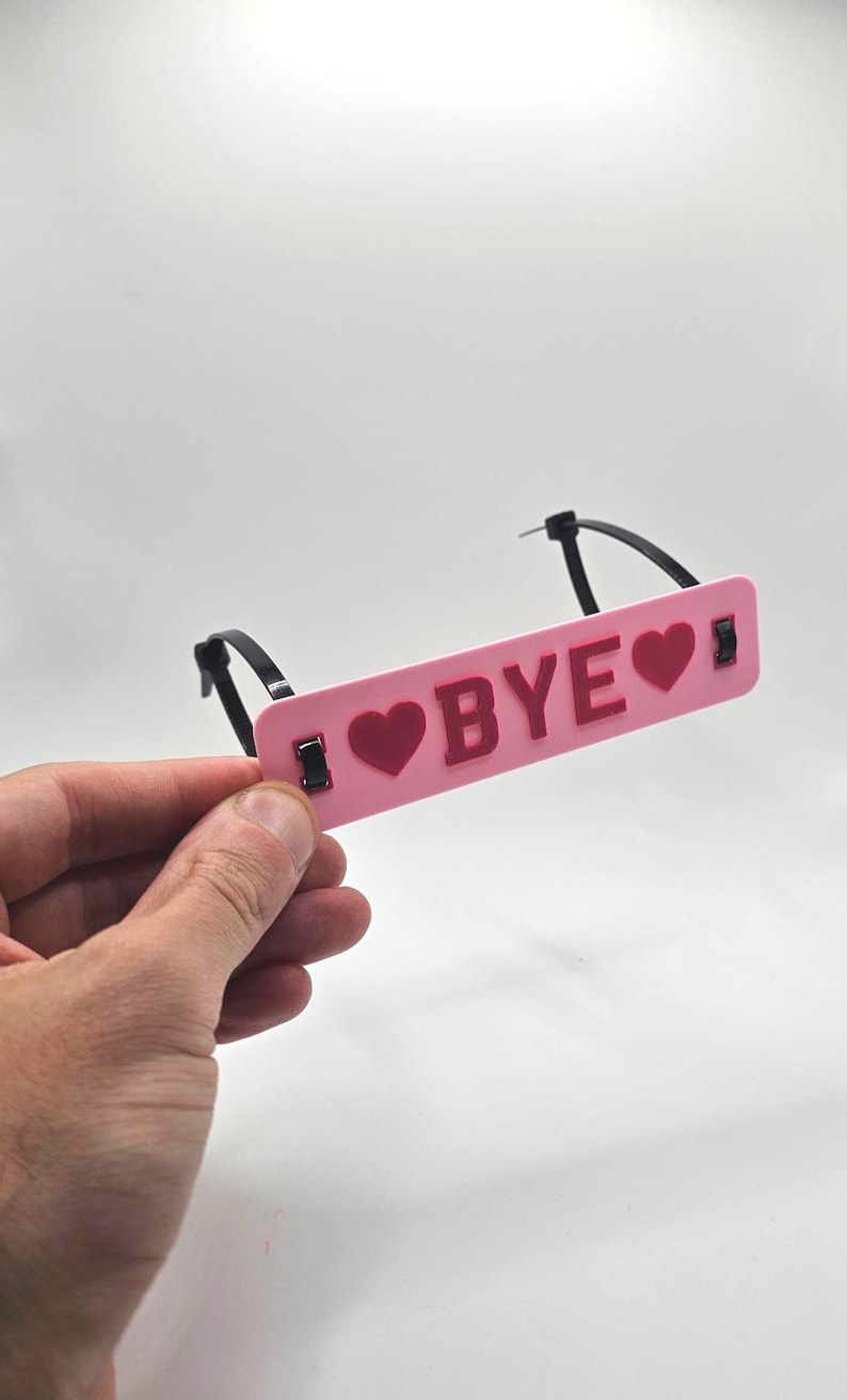 May include: A pink luggage tag with the word "BYE" and heart symbols in dark pink. The tag is held by black zip ties. The background is plain white.