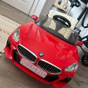 May include: A bright red toy car with a black grill and the BMW logo. The car has a windshield, side mirrors, and a steering wheel. The name "NELLIE RAE" is displayed on a pink license plate.