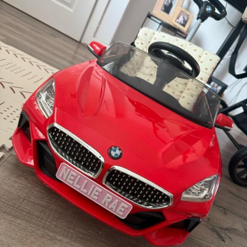 May include: A bright red toy car with a black grill and the BMW logo. The car has a windshield, side mirrors, and a steering wheel. The name "NELLIE RAE" is displayed on a pink license plate.
