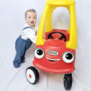 May include: A red and yellow toy car with the name "Rosie" on the front. The car has large, cartoonish eyes and a smiling face. The car has black wheels with white accents. A child is standing next to the car.