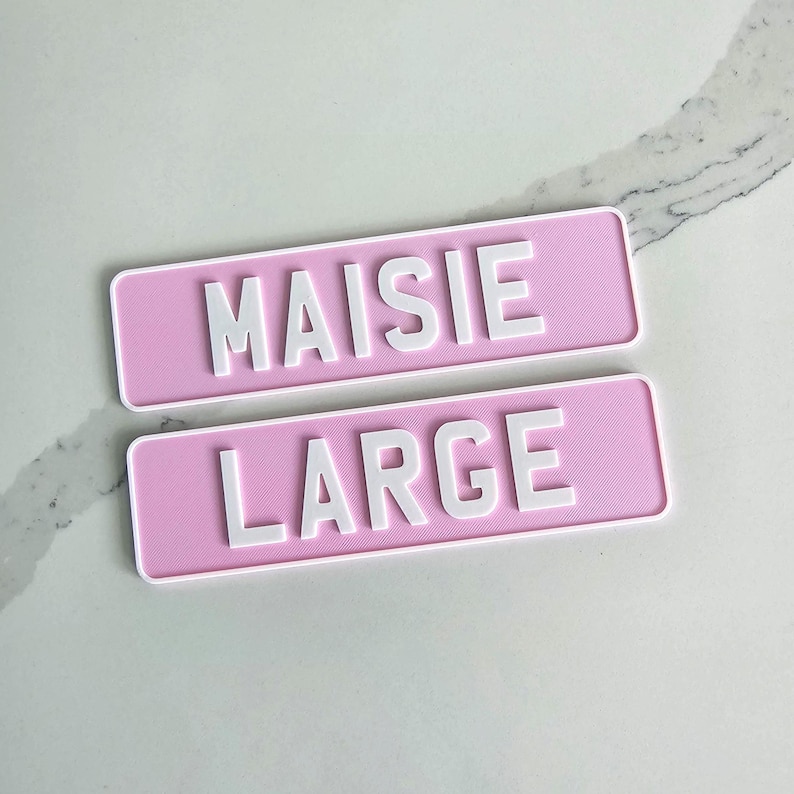 May include: Two rectangular pink signs with white text. The top sign reads "MAISIE" and the bottom sign reads "LARGE". The signs have a textured surface and a white border.