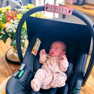 May include: A navy blue baby car seat with a pink sign that reads "DA BABY". The car seat has a baby wearing a floral print onesie. A bouquet of flowers is in the background.