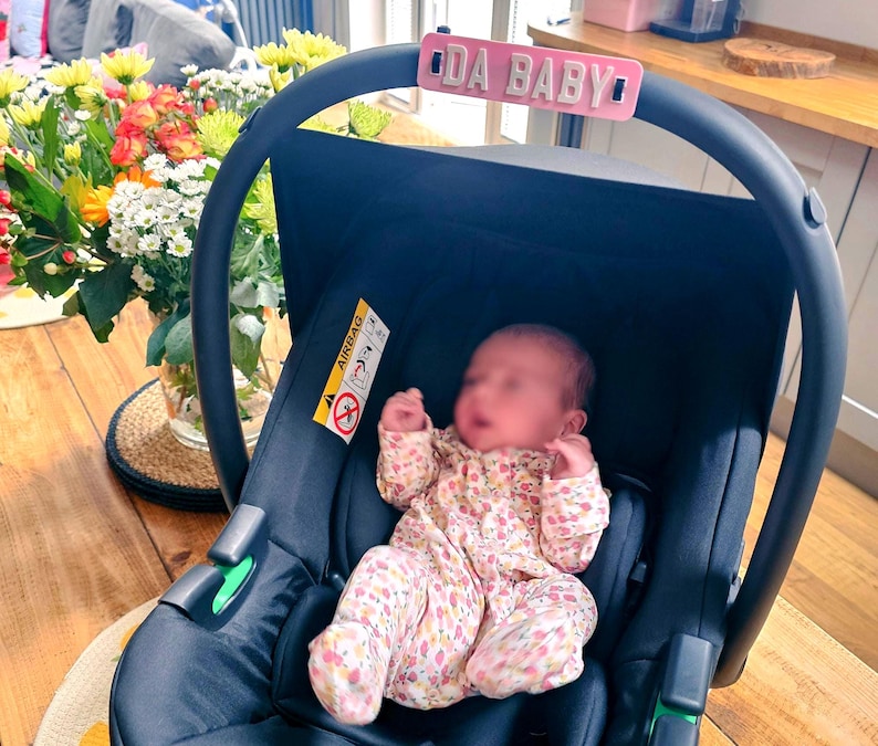 May include: A navy blue baby car seat with a pink sign that reads "DA BABY". The car seat has a baby wearing a floral print onesie. A bouquet of flowers is in the background.