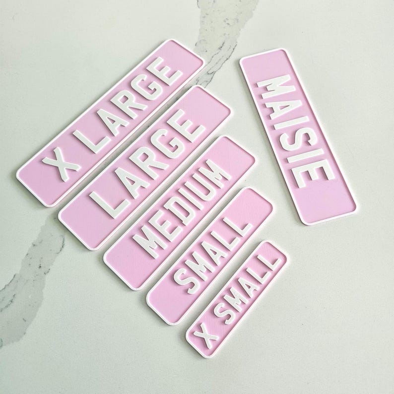 May include: A collection of pink rectangular labels with white raised lettering. The labels display the words "X LARGE", "LARGE", "MEDIUM", "SMALL", and "X SMALL". A separate label reads "MAISIE".