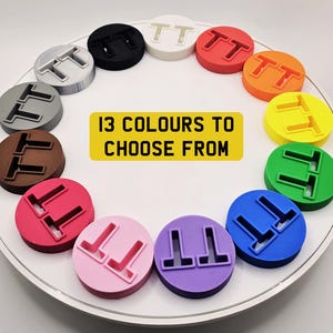 May include: A collection of 13 colorful, circular objects, each with a raised "TT" design. The objects are arranged in a circle on a white surface. A yellow rectangle in the center reads "13 COLOURS TO CHOOSE FROM".