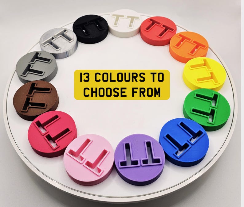 May include: A collection of 13 colorful, circular objects, each with a raised "TT" design. The objects are arranged in a circle on a white surface. A yellow rectangle in the center reads "13 COLOURS TO CHOOSE FROM".