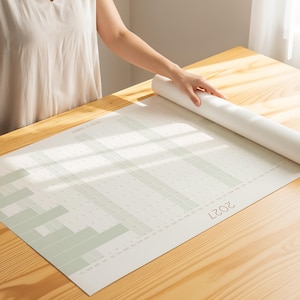Peut inclure: Un planificateur annuel blanc pour 2027 avec une grille et des accents vert pâle. Le planificateur est déroulé sur une surface en bois. Une partie enroulée du planificateur est visible, avec une main.