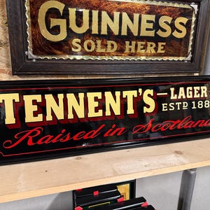 May include: A black framed sign with the text "TENNENT'S - LAGER - ESTD 1885 Raised in Scotland" in gold and red lettering. A "Guinness SOLD HERE" sign is visible above it.