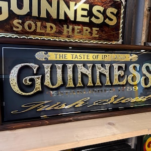 Peut inclure: Un miroir vintage encadré noir et or avec le texte "The Taste of Ireland Guinness Established 1759 Irish Stout" sur un fond sombre.