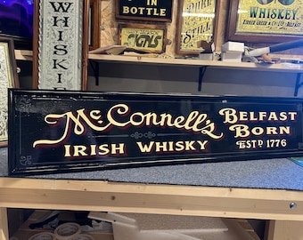 Könnte beinhalten: Schwarzes Schild mit goldener Beschriftung, das für McConnells Irish Whisky wirbt. Das Schild lautet "McConnells Belfast Born Irish Whisky Est. 1776".