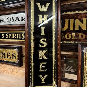 Può includere: Un'insegna rettangolare in legno con la scritta "WHISKEY" in lettere dorate su sfondo nero. L'insegna ha una cornice marrone scuro con finiture dorate ed è esposta con altre insegne di bar.