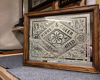 Könnte beinhalten: Ein gerahmter Spiegel mit silbernem Hintergrund und einem schwarzen und weißen rautenförmigen Logo, auf dem "Kirker & Greer Whiskey Limited" steht, mit einem Blumendesign in der Mitte. Das Logo ist von einem dekorativen Rahmen umgeben.
