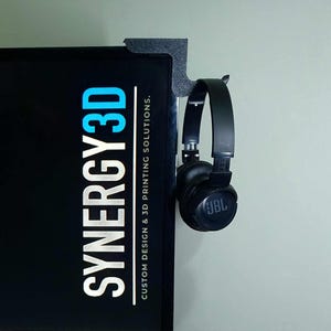 May include: A black 3D printed headphone holder attached to a black surface with the text "SYNERGY3D CUSTOM DESIGN & 3D PRINTING SOLUTIONS." A pair of black JBL headphones are hanging on the holder.