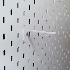May include: White plastic pegboard hook with a curved arm. The hook is mounted on a white pegboard with a grid of evenly spaced holes.
