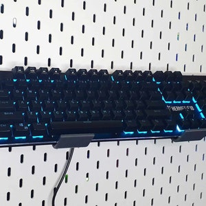 May include: A black keyboard with blue backlighting is mounted on a white pegboard wall. The keyboard has the text "THERMALTAKE" printed on the right side.