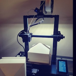 May include: A dark blue 3D printer with a white filament spool and a touchscreen display. The printer is in operation, creating a white geometric form. The brand name "ELEGOO" is visible on the machine.