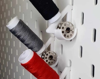 May include: Three white plastic spool holders with metal bobbins, mounted on a white pegboard wall. The spools are filled with black, gray, and red thread.
