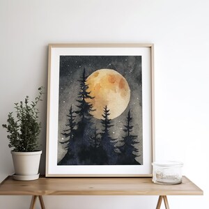 May include: Watercolor painting of a full moon over a silhouetted forest. The moon is a warm yellow-orange, and the trees are dark blue-black. The artwork is framed in a light wood frame and is displayed on a wooden surface.