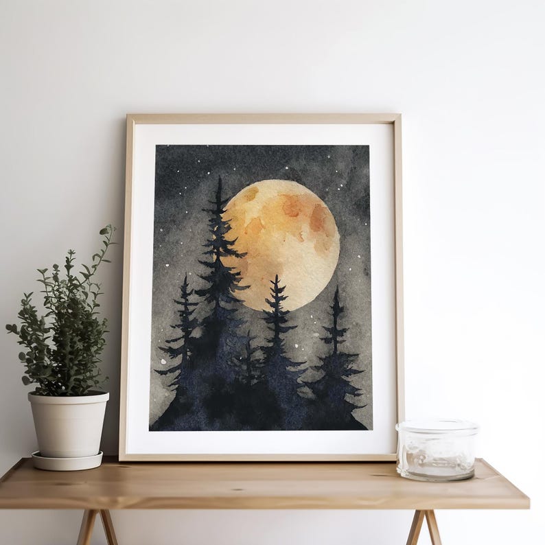 May include: Watercolor painting of a full moon over a silhouetted forest. The moon is a warm yellow-orange, and the trees are dark blue-black. The artwork is framed in a light wood frame and is displayed on a wooden surface.