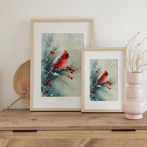 Op de afbeelding: Twee ingelijste aquarelprints van een rode kardinaal die op een besneeuwde tak met rode bessen zit. De prints worden tentoongesteld op een houten dressoir met een decoratieve waaier en een vaas met gedroogde bloemen.