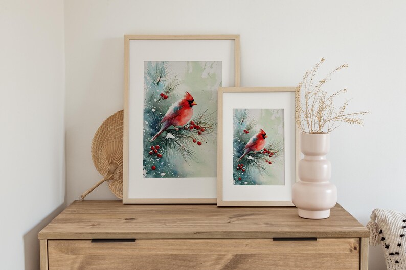 Op de afbeelding: Twee ingelijste aquarelprints van een rode kardinaal die op een besneeuwde tak met rode bessen zit. De prints worden tentoongesteld op een houten dressoir met een decoratieve waaier en een vaas met gedroogde bloemen.