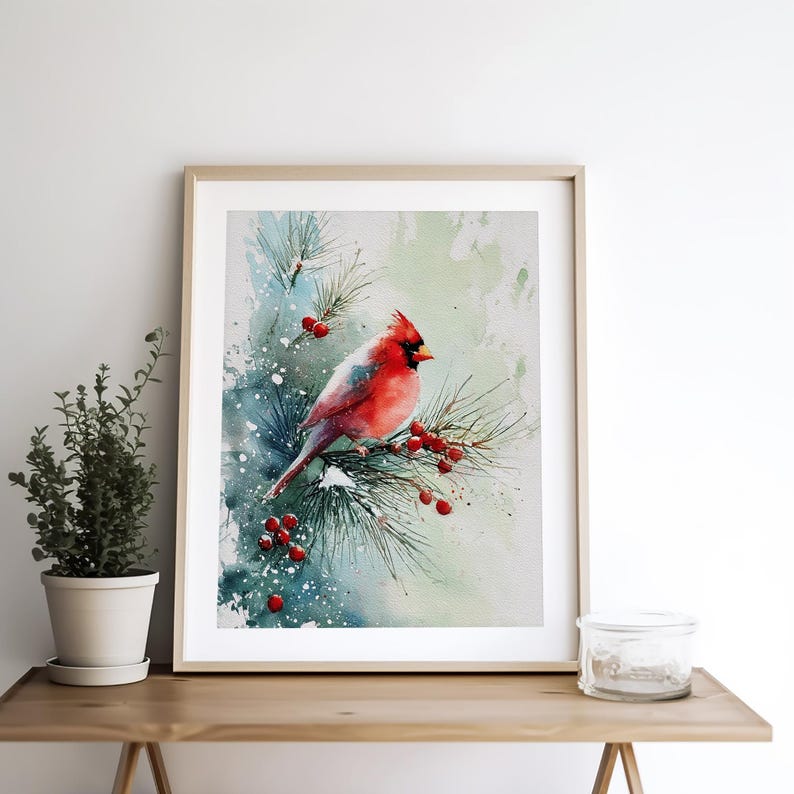 Op de afbeelding: Aquarel schilderij van een rode kardinaal die op een met sneeuw bedekte dennentak met rode bessen zit. Het kunstwerk is ingelijst in een licht houten frame en tegen een zachte aquarel achtergrond.
