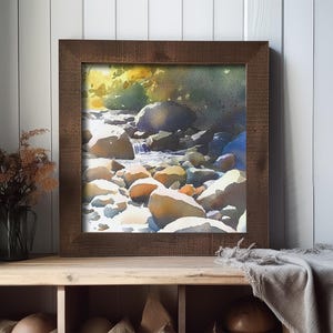 May include: A framed watercolor painting of a stream flowing over rocks in a forest. The painting is in a brown wooden frame and is hanging on a white wall.