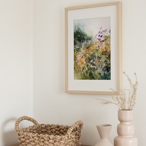 May include: A framed watercolor painting of a field of wildflowers in shades of purple, yellow, and green. The painting is hanging on a white wall above a wooden dresser with a woven basket and two ceramic vases.