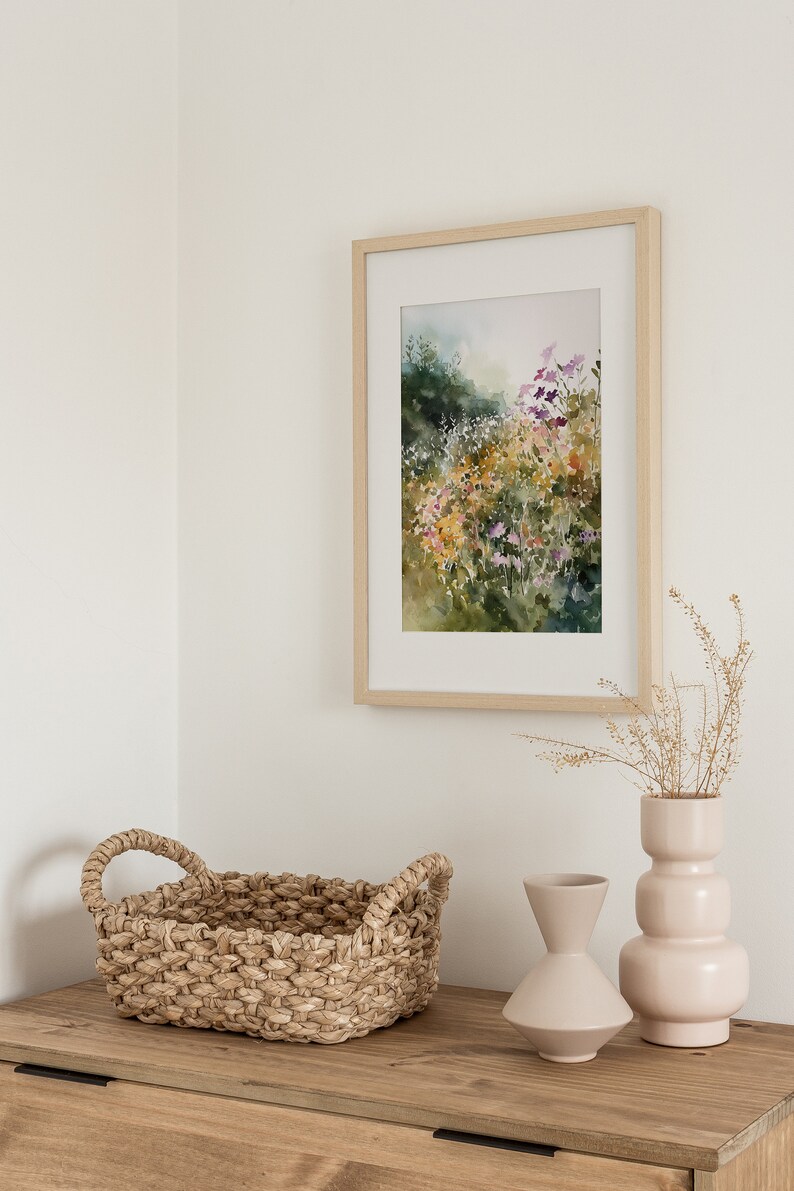 May include: A framed watercolor painting of a field of wildflowers in shades of purple, yellow, and green. The painting is hanging on a white wall above a wooden dresser with a woven basket and two ceramic vases.