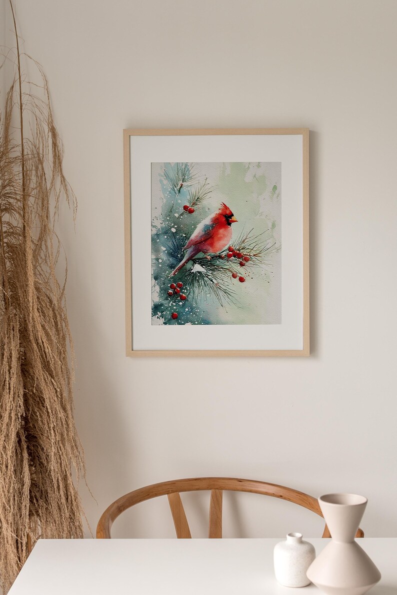 Op de afbeelding: Waterverfschilderij van een rode kardinaal die op een met sneeuw bedekte dennentak met rode bessen zit. Het kunstwerk is ingelijst in een licht houten frame, waardoor een serene winterse sc&egrave;ne ontstaat. Het schilderij wordt in een goed verlichte ruimte tentoongesteld.
