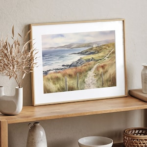 May include: A framed watercolour painting of a coastal landscape with a path leading to a small white house. The painting features a blue ocean with white waves, green grass, and a rocky coastline.