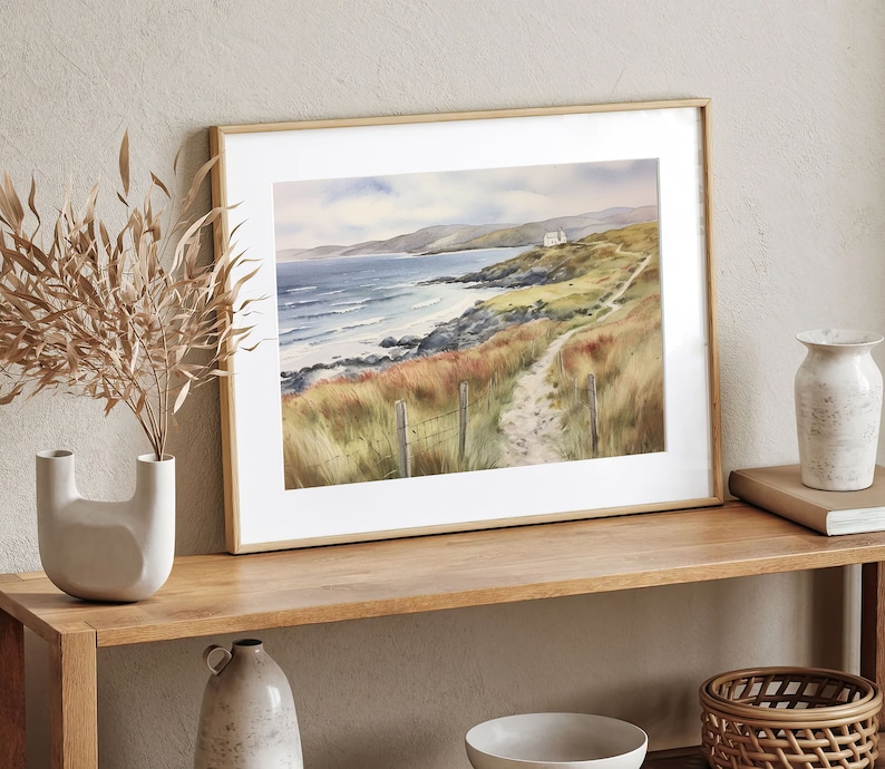 May include: A framed watercolour painting of a coastal landscape with a path leading to a small white house. The painting features a blue ocean with white waves, green grass, and a rocky coastline.
