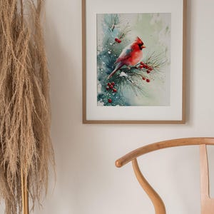 Op de afbeelding: Waterverfschilderij van een rode kardinaal die op een dennentak met rode bessen zit. Het kunstwerk is ingelijst in een lichtbruine houten lijst en hangt aan een witte muur. Het schilderij heeft een winterthema.