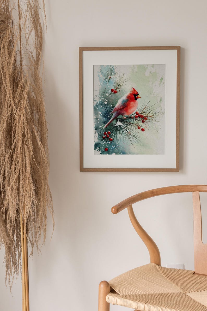 Op de afbeelding: Waterverfschilderij van een rode kardinaal die op een dennentak met rode bessen zit. Het kunstwerk is ingelijst in een lichtbruine houten lijst en hangt aan een witte muur. Het schilderij heeft een winterthema.