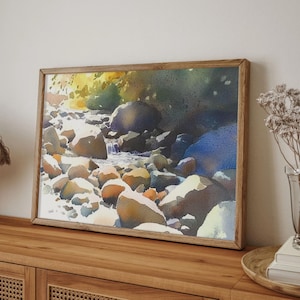 May include: A framed watercolor painting of a stream flowing through a rocky landscape. The water is a light blue, and the rocks are a variety of browns and grays. The background is a dark green.