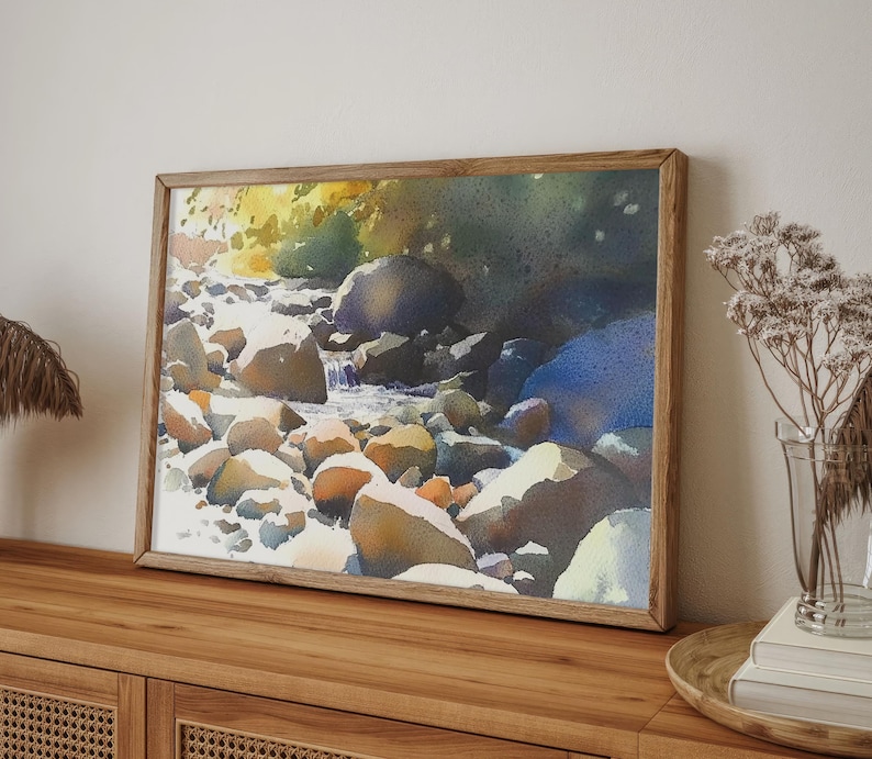 May include: A framed watercolor painting of a stream flowing through a rocky landscape. The water is a light blue, and the rocks are a variety of browns and grays. The background is a dark green.