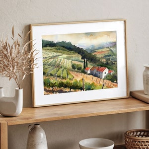May include: Watercolor painting of a vineyard landscape with a white house and red roof. The artwork is framed in a light wood frame and depicts rows of grapevines, rolling hills, and a cloudy sky. The overall color palette is green, brown, and beige.