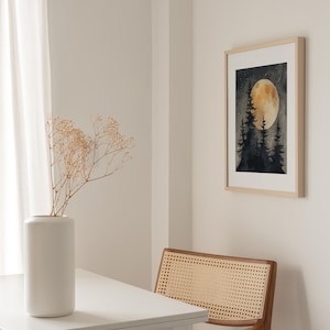 May include: A framed watercolor print depicts a nighttime forest scene with a large, glowing moon. The artwork hangs on a white wall, next to a wooden chair with a woven back. A white vase with dried flowers sits on a white table.