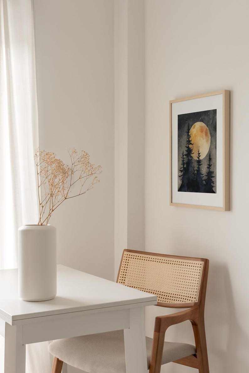 May include: A framed watercolor print depicts a nighttime forest scene with a large, glowing moon. The artwork hangs on a white wall, next to a wooden chair with a woven back. A white vase with dried flowers sits on a white table.