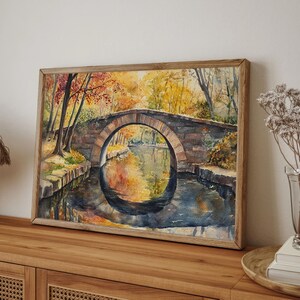 May include: Watercolor painting of a stone bridge over a calm river, framed in a light wood. The artwork depicts an autumn scene with trees in shades of red, orange, and yellow reflected in the water. The bridge has an arched design.