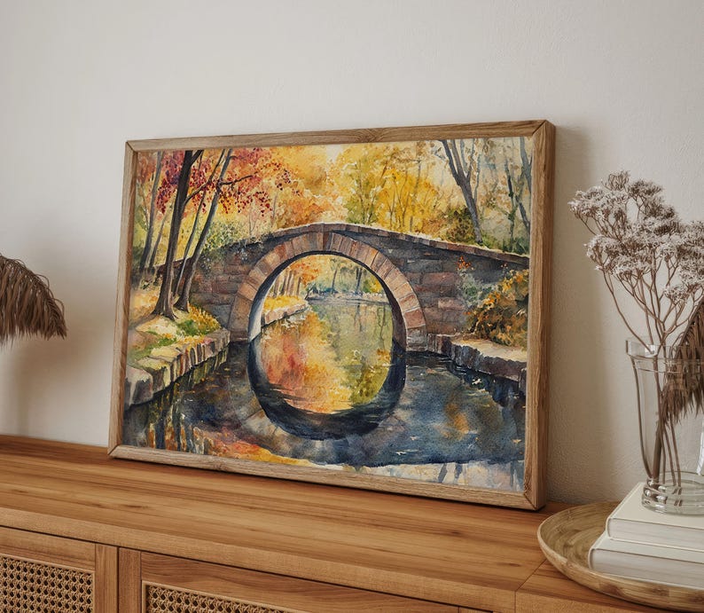 May include: Watercolor painting of a stone bridge over a calm river, framed in a light wood. The artwork depicts an autumn scene with trees in shades of red, orange, and yellow reflected in the water. The bridge has an arched design.