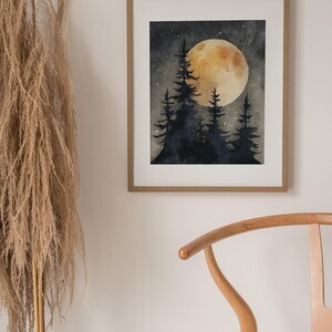 May include: A framed watercolor painting depicts a nighttime forest scene. A large, yellow moon glows behind silhouetted pine trees. The artwork is displayed in a light brown wooden frame, with a neutral-toned wall and a wooden chair in the background.
