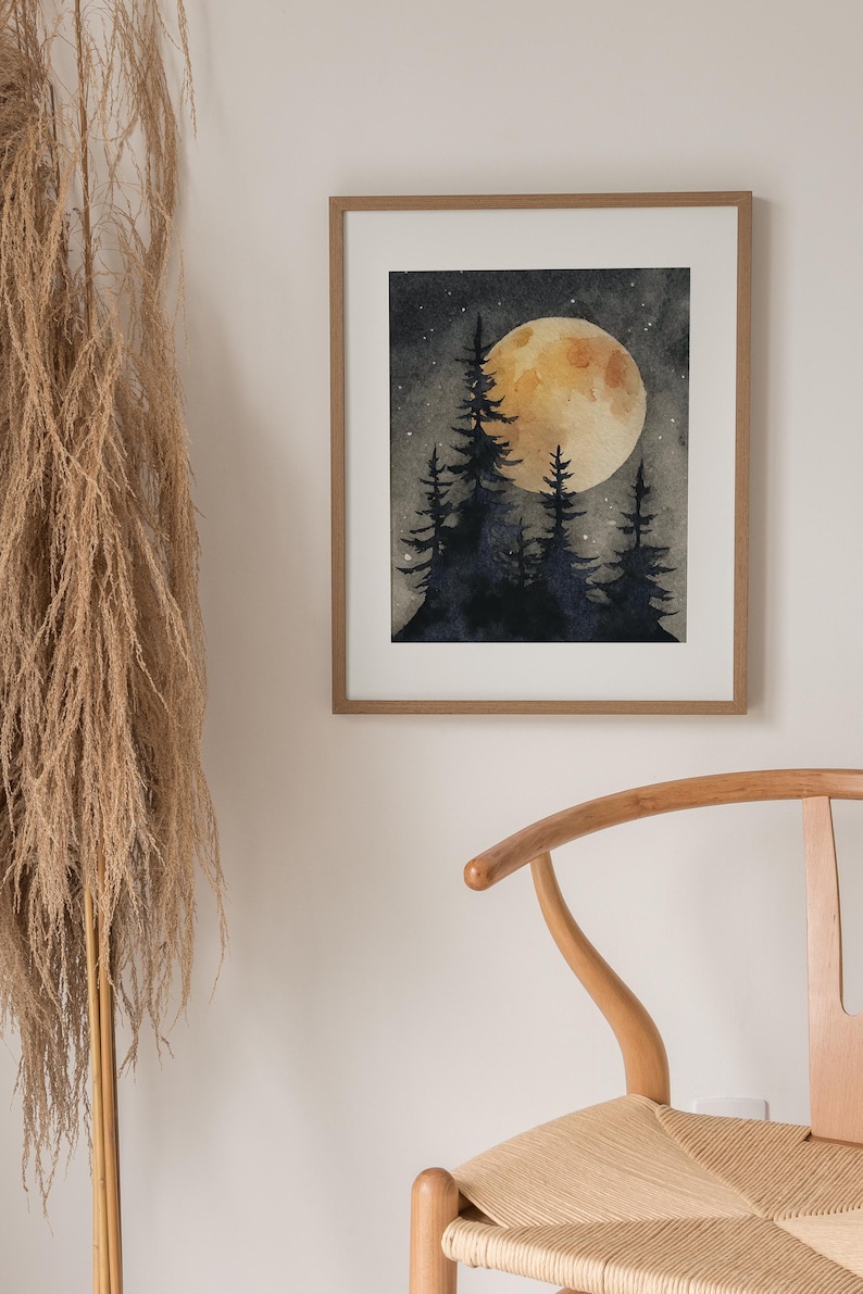 May include: A framed watercolor painting depicts a nighttime forest scene. A large, yellow moon glows behind silhouetted pine trees. The artwork is displayed in a light brown wooden frame, with a neutral-toned wall and a wooden chair in the background.