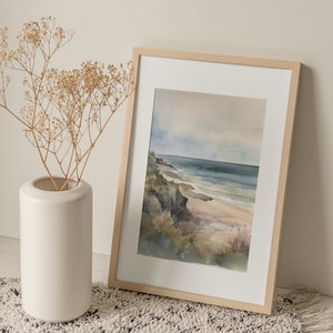 May include: A framed watercolor painting of a seascape with a sandy beach, rocky cliffs, and blue water. The painting is in a light wood frame and is sitting on a white fluffy rug.