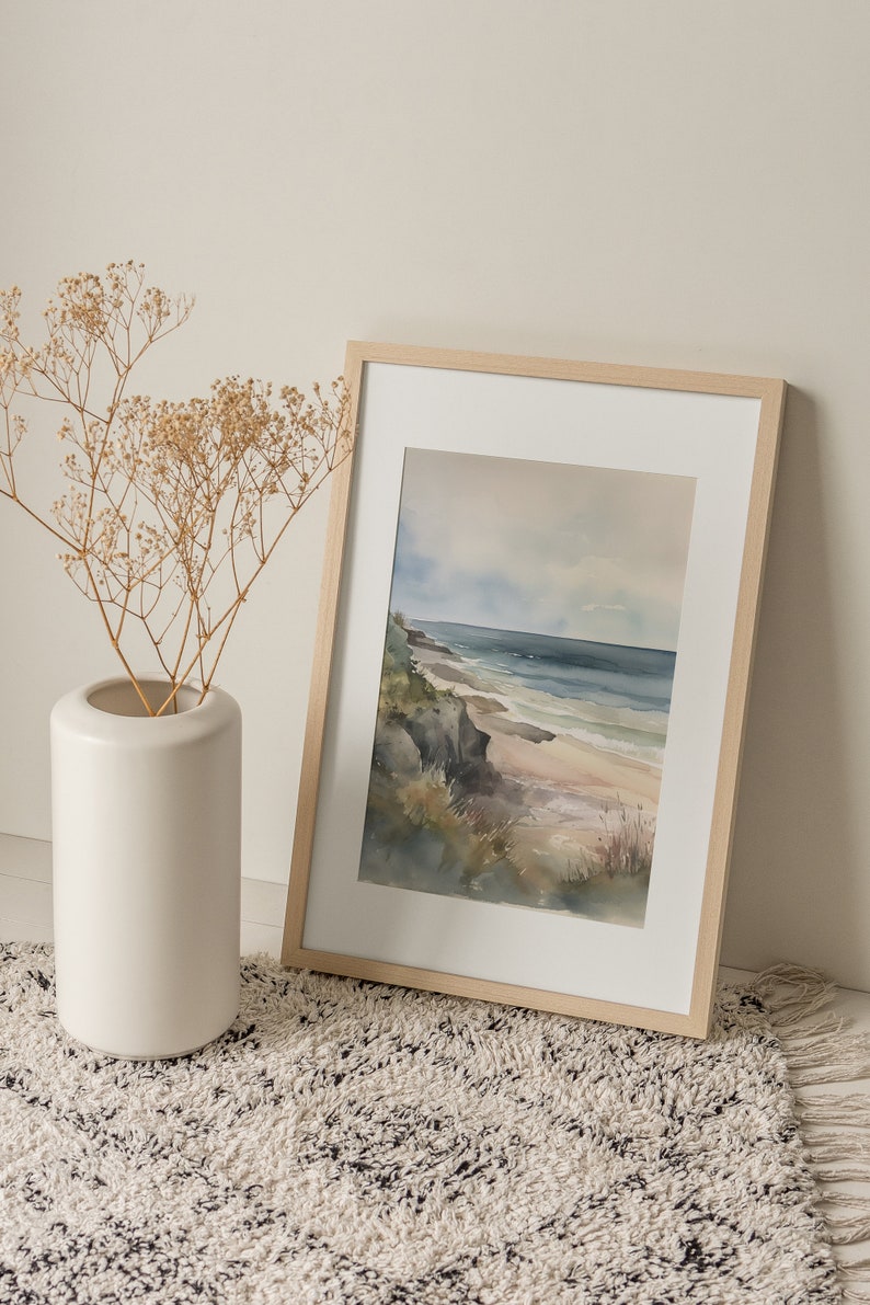 May include: A framed watercolor painting of a seascape with a sandy beach, rocky cliffs, and blue water. The painting is in a light wood frame and is sitting on a white fluffy rug.